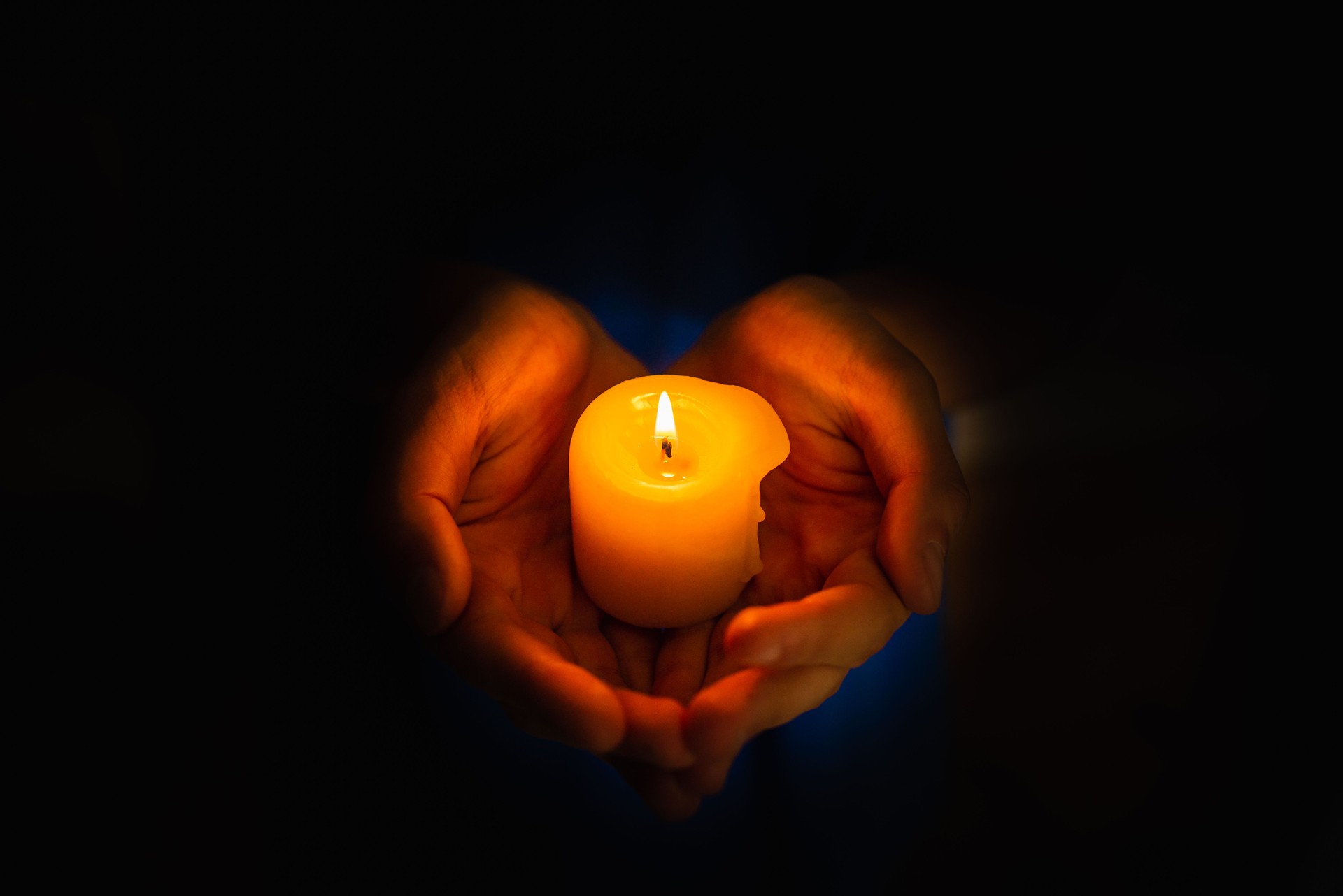 Hands holding burning candle in darkness like a heart, black background.